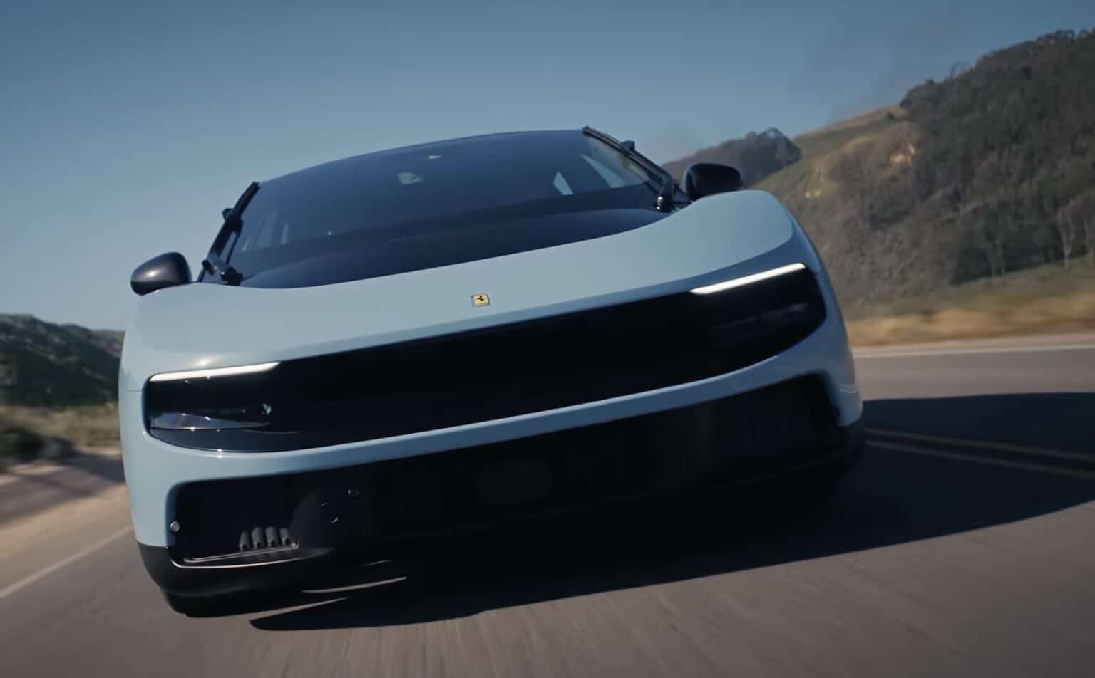 Hamilton and Leclerc rijden de Ferrari Luce Light blue sports car speeding toward camera on a highway, windshield wipers visible and hills in the background.